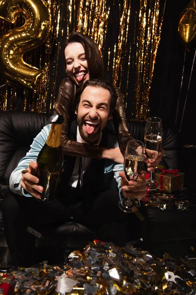 Fröhliches Paar streckt die Zungen aus, während es das Jahr mit Champagner auf Schwarz feiert — Stockfoto