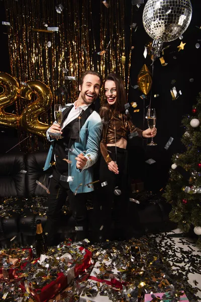 Pareja alegre y elegante con bengalas celebrando la Navidad cerca de la decoración brillante en negro - foto de stock