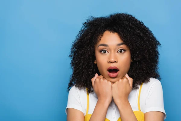 Mulher afro-americana surpreendida segurando punhos perto do queixo isolado em azul — Fotografia de Stock