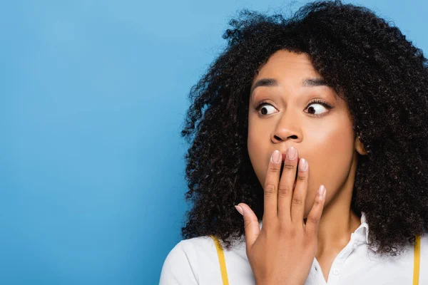 Chocado afro-americano mulher cobrindo boca com a mão enquanto olhando para longe isolado no azul — Fotografia de Stock