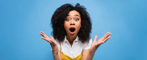 Femme afro-américaine étonnante montrant wow geste tout en regardant la caméra isolée sur bleu, bannière — Photo de stock