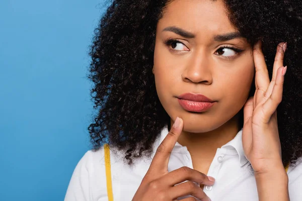 Reflexiva mujer afroamericana tocando la barbilla mientras mira hacia otro lado aislado en azul - foto de stock