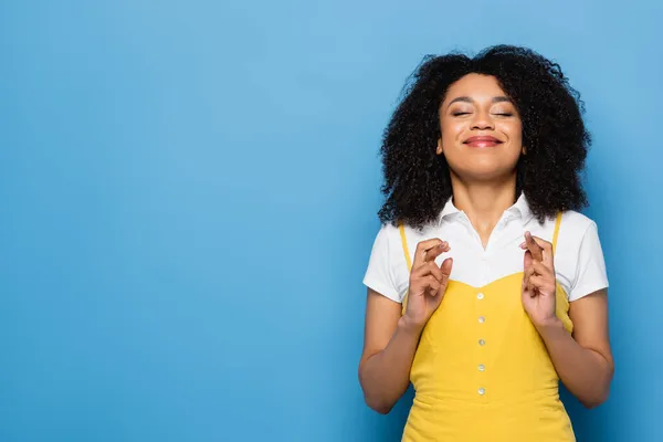 Femme afro-américaine souriante avec les yeux fermés tenant les doigts croisés pour la chance sur bleu — Photo de stock