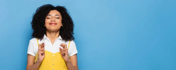 Mujer afroamericana positiva con los ojos cerrados sosteniendo los dedos cruzados aislados en azul, pancarta - foto de stock