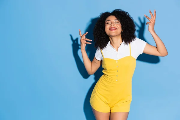 Glückliche afrikanisch-amerikanische Frau mit geschlossenen Augen gestikuliert, während sie auf blau posiert — Stockfoto