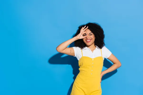 Fröhliche afrikanisch-amerikanische Frau zeigt okay Zeichen in der Nähe Gesicht, während sie mit der Hand auf der Hüfte auf blau posiert — Stockfoto