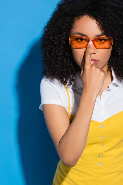 Afro-américaine femme regardant caméra tout en ajustant les lunettes de vue à la mode sur bleu — Photo de stock