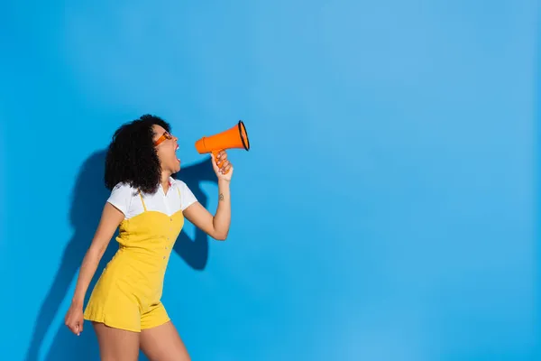African american woman in yellow jumpsuit shouting in megaphone on blue — Stock Photo