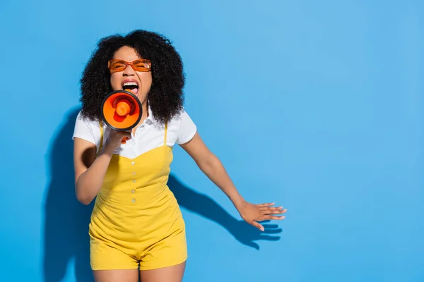 African american woman in orange eyeglasses screaming in megaphone on blue — Stock Photo