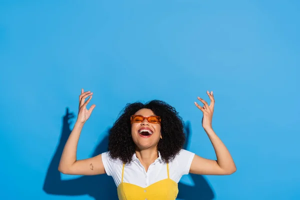 Excited african american woman in stylish eyeglasses laughing with closed eyes on blue — Stock Photo