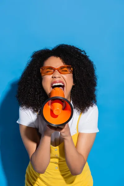 Thrilled african american woman in orange eyeglasses screaming in megaphone on blue — Stock Photo