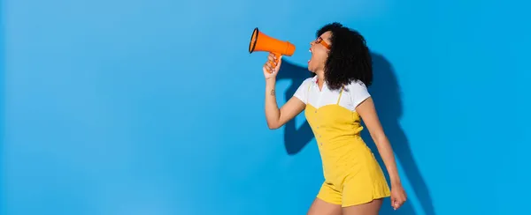 Energized african american woman in trendy jumpsuit screaming in megaphone on blue, banner — Stock Photo