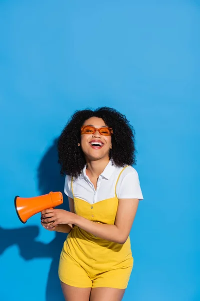 Femme afro-américaine gaie dans des lunettes à la mode tenant mégaphone sur bleu — Photo de stock