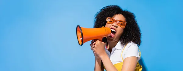 Excited african american woman in eyeglasses shouting in loudspeaker on blue, banner — Stock Photo