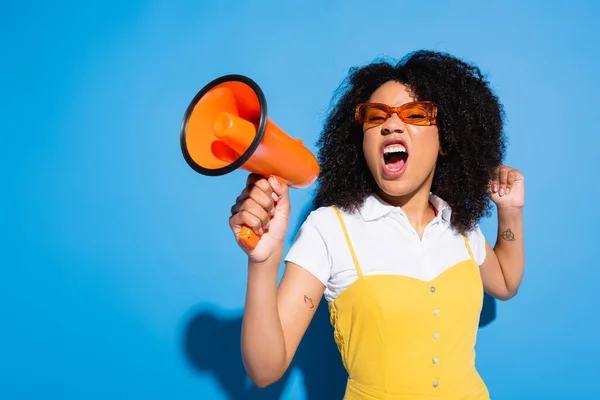 Aufgeregte afrikanisch-amerikanische Frau mit Brille schreit in orangefarbenem Lautsprecher auf blau — Stockfoto