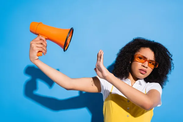 Donna afroamericana scontenta in occhiali alla moda mostrando gesto stop vicino megafono su blu — Foto stock