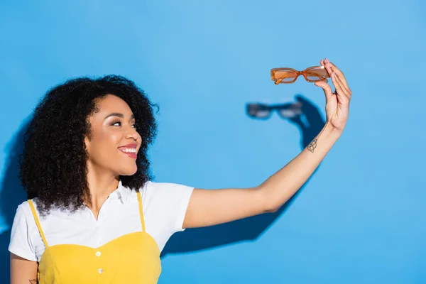 Cheerful african american woman holding trendy eyeglasses on blue — Stock Photo