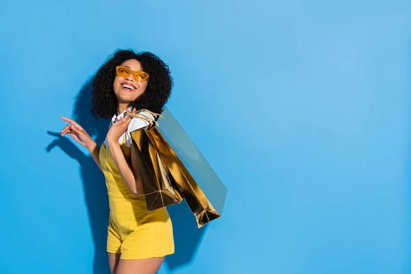 Збуджена афроамериканка в жовтих окулярах позує з блискучими сумками на синьому — стокове фото