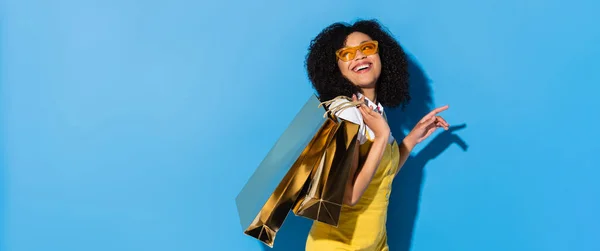 Alegre y elegante mujer afroamericana con bolsas de compras brillantes mirando hacia otro lado en azul, bandera - foto de stock