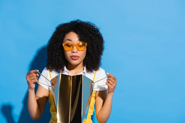 Surpris femme afro-américaine en lunettes jaunes regardant dans le sac à provisions brillant sur bleu — Photo de stock