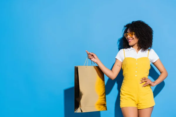 Mujer afroamericana positiva en mono amarillo posando con la mano en la cadera y bolsa de compras brillante en azul - foto de stock