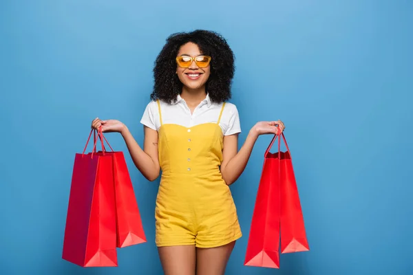 Счастливая африканская американка в желтых очках и комбинезоне позирует с пакетами покупок на голубом — стоковое фото