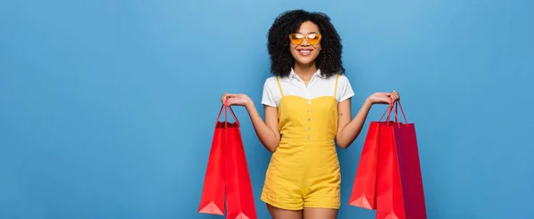 Cheerful african american woman in yellow jumpsuit and eyeglasses standing with shopping bags isolated on blue, banner — Stock Photo