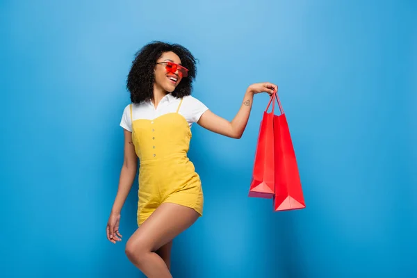 Trendy african american woman in red eyeglasses posing with coral shopping bags on blue — Stock Photo
