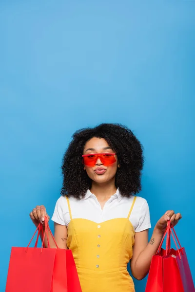 Trendy african american woman with coral shopping bags pouting lips isolated on blue — Stock Photo