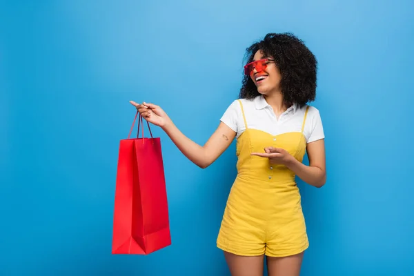 Fröhliche afrikanisch-amerikanische Frau in roter Brille und gelbem Overall, die auf Korallen-Einkaufstasche auf blau zeigt — Stockfoto