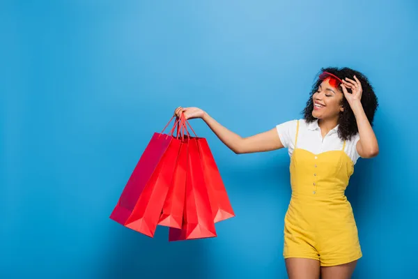 Joyful african american woman in trendy jumpsuit and eyeglasses holding coral shopping bags on blue — Stock Photo