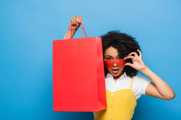 African american woman touching stylish eyeglasses while showing coral shopping bag on blue — Stock Photo