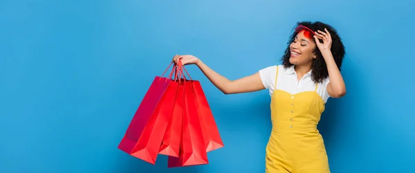 Cheerful african american woman in yellow jumpsuit holding coral shopping bags on blue, banner — Stock Photo