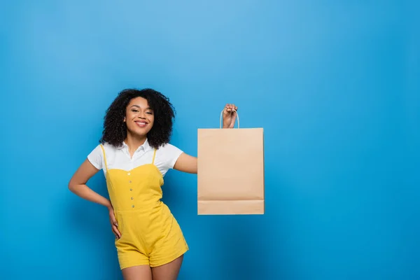 Fröhliche afrikanisch-amerikanische Frau im gelben Overall posiert mit der Hand auf der Hüfte und der Einkaufstasche auf blau — Stockfoto