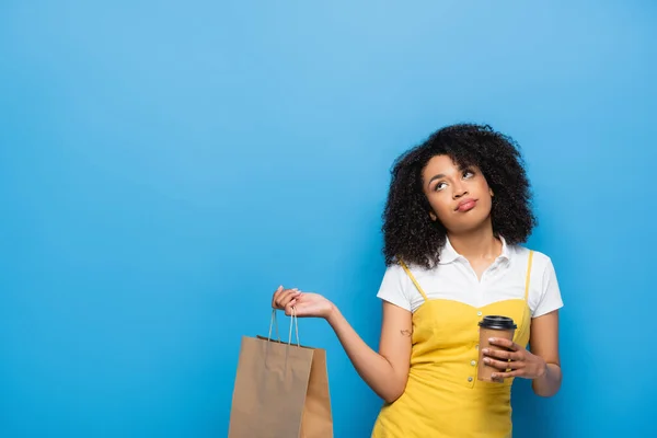 Mulher afro-americana descontente com copo de papel e saco de compras olhando para cima em azul — Fotografia de Stock