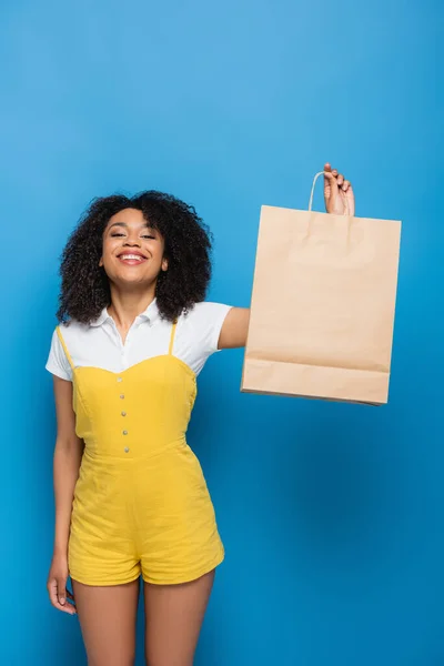 Mulher americana africana satisfeito em macacão amarelo segurando saco de compras enquanto olha para a câmera em azul — Fotografia de Stock