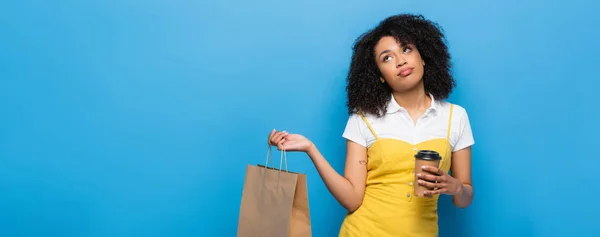 Mujer afroamericana molesta con café para ir y bolsa de compras mirando hacia arriba en azul, pancarta - foto de stock