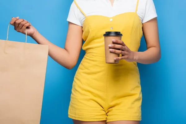 Частичный вид на африканскую американку в жёлтом комбинезоне с пакетом для покупок и бумажной чашкой на синем — стоковое фото