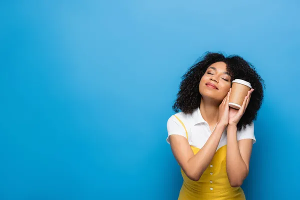 Pleased african american woman with closed eyes holding disposable cup on blue — Stock Photo