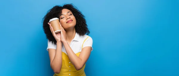 Mujer afroamericana feliz con los ojos cerrados sosteniendo café para ir en azul, bandera - foto de stock