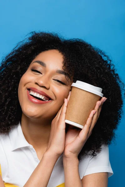 Joyeuse femme afro-américaine tenant tasse en papier près du visage isolé sur bleu — Photo de stock