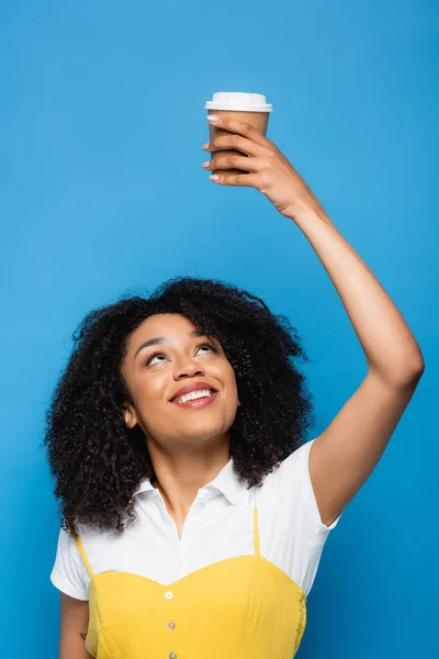 Smiling african american woman holding takeaway drink in raised hand isolated on blue — Stock Photo