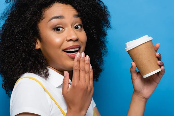 Étonnante femme afro-américaine avec du café pour aller couvrir la bouche avec la main isolée sur bleu — Photo de stock