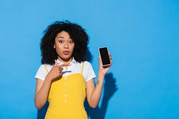 Überrascht afrikanisch-amerikanische Frau blickt in Kamera, während sie auf Handy mit leerem Bildschirm auf blau zeigt — Stockfoto