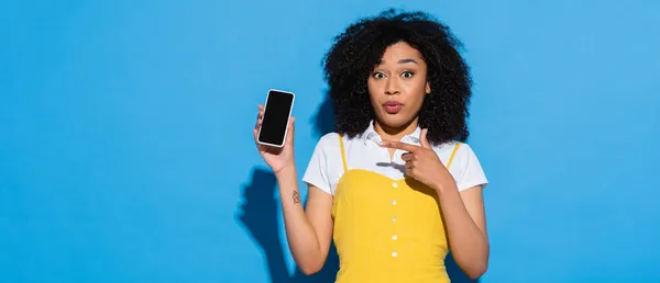 Mulher americana africana espantado apontando para o telefone móvel com tela em branco no azul, banner — Fotografia de Stock