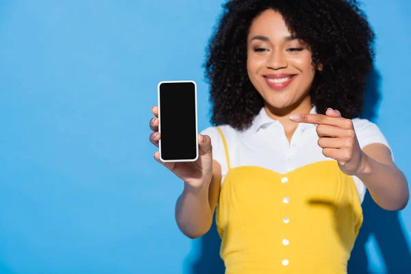 Mujer afroamericana borrosa apuntando al teléfono móvil con pantalla en blanco en azul - foto de stock