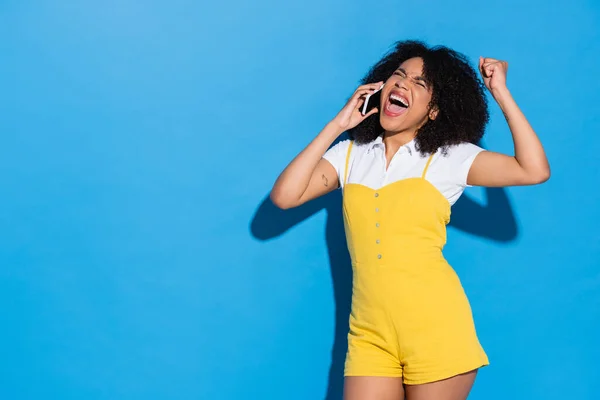 Mujer afroamericana emocionada en mono amarillo mostrando gesto de victoria mientras habla en el teléfono celular en azul - foto de stock