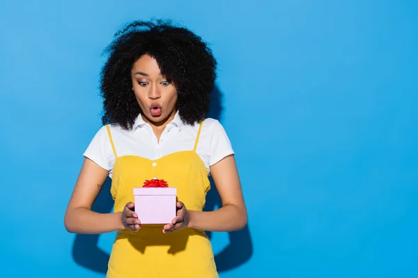 Amazed african american woman with open mouth holding gift box on blue — Stock Photo
