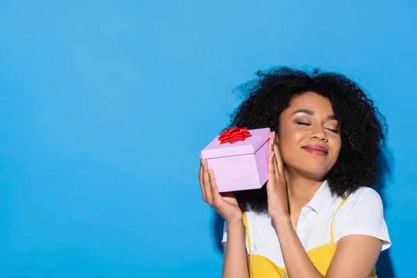 Erfreut afrikanisch-amerikanische Frau mit geschlossenen Augen hält Geschenk auf blau — Stockfoto