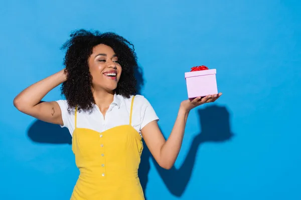 Fröhliche afrikanisch-amerikanische Frau hält Geschenkbox mit roter Schleife auf blauem Grund — Stockfoto